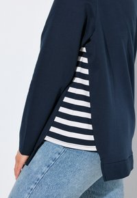 Marineblaue Langarmoberteil mit seitlichen Schlitzen, über einem unterteil in Weiß und Marineblau mit Streifen. Baumwollmischung mit glatter Textur.