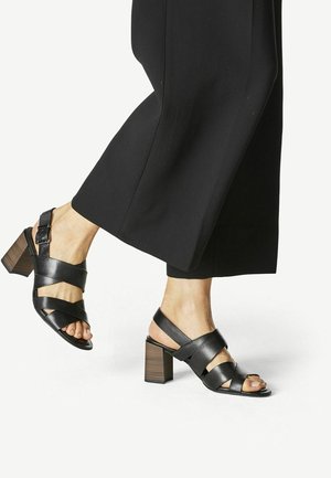 Jambe portant un pantalon large noir et des sandales noires en cuir à talon bloc avec talons à texture boisée sur fond blanc.