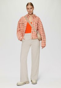 Geweven oranje en room houndstooth jas, gecombineerd met een feloranje top, beige broek en roomkleurige schoenen met een dikke zool. Kleine beige tas.