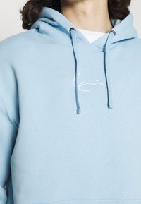 Light blue hoodie made of soft fabric, featuring a front kangaroo pocket, adjustable drawstrings, and a small white embroidered logo.