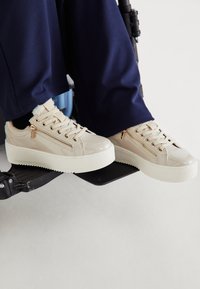 Μπεζ γυαλιστερά δερμάτινα αθλητικά παπούτσια με ενισχυμένη σόλα, που διαθέτουν διακοσμητικούς φερμουάρ, σχέδιο με κορδόνια και ανοιχτόχρωμες λεπτομέρειες.