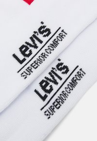 Bele bombažne nogavice z črnim logotipom "Levi's" in napisom "vrhunska udobnost", ki prikazujejo mehak pleteni vzorec in kontrastne barve.
