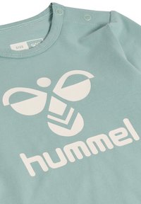 Ljus teal bomullströja med en vit grafisk logotyp och texten "hummel." Har tryckknappar vid axeln för enkel påklädning.