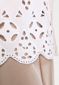 Vit broderad spetskant med utklippta mönster ovanpå en beige tygbakgrund, med vågiga kanter och intrikata detaljer.