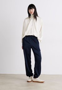 Lysebeige oversized zip-up hettegenser med elastiske mansjetter, parret med marineblå smale bukser og hvite joggesko med brune såler. Glatt tekstur.
