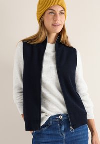 Marineblauer Strickweste mit Reißverschluss, getragen über einem strukturierten cremefarbenen Pullover. Das Model trägt eine gelbe Rippmütze. Darunter sind Jeans aus Denim sichtbar.