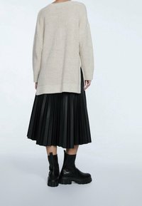 Beige gebreide trui met zijsplitten, gecombineerd met een zwarte plissérok en chunky zwarte enkellaarsjes, met gestructureerde materialen en gelaagd ontwerp.