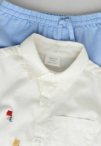 Chemise blanche à boutons avec des détails en broderie, associée à un short bleu clair avec une taille élastique. Texture de tissu lisse visible.