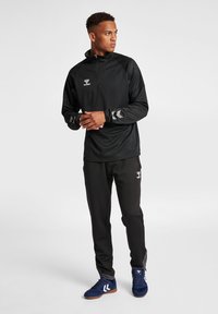 Pullover atletico nero con colletto alto e zip parziale, abbinato a pantaloni neri. Presenta un tessuto testurizzato e accenti a contrasto.