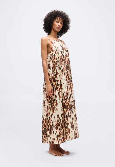 Γυναίκα με σγουρά μαλλιά, όρθια, ξυπόλυτη, φοράει αμάνικο φόρεμα μέχρι τον αστράγαλο με καφέ και μαύρο πουά μοτίβο σε κρεμ ύφασμα.