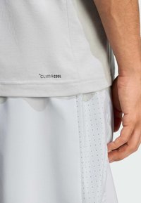 Camiseta deportiva de color gris claro con la marca «CLIMACOOL» en el dobladillo, combinada con pantalones cortos blancos con pequeñas perforaciones. Textura de tela suave.