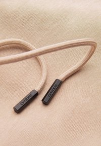 Beige geflochtene Kordel mit schwarzen Kunststoffspitzen, geprägt mit "ombre", liegt auf passendem strukturiertem Stoff.