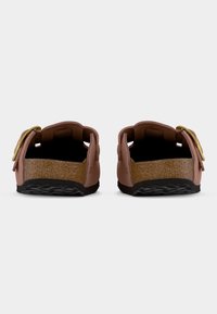 Ζευγάρι ροζ σαγιονάρες χωρίς λουράκια με φελλό στη σόλα, μαύρες σόλες και ρυθμιζόμενες χρυσές αγκράφες, απεικονισμένες από πίσω με λευκό φόντο.