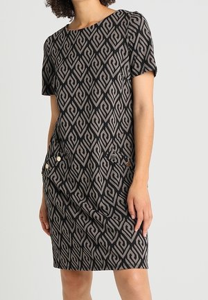 Kobieta w czarnej sukience z krótkim rękawem, sięgającej do kolan, z szarym geometrycznym wzorem i dwoma zapinanymi na guziki kieszeniami z przodu.