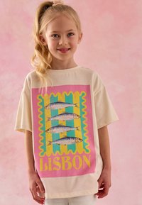 Krém színű póló, amely élénk grafikával díszített, szardíniákkal egy rózsaszín, sárga és türkiz háttéren. A szöveg így szól: „LISZABON.”