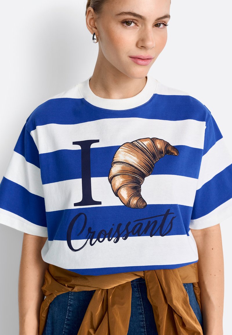 Femme portant une chemise rayée bleue et blanche avec le texte "J'aime les croissants" (avec une image de croissant), assortie à un jean et une veste marron nouée à la taille.