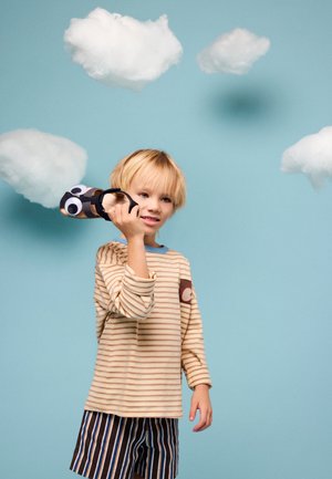 Niño rubio joven sosteniendo un avión de juguete con ojos móviles sobre un fondo azul con nubes blancas flotantes.