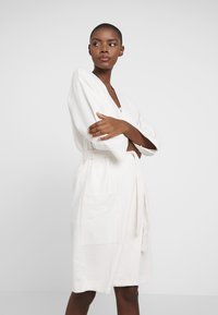 Wafeltextuur, knielange witte jurk met V-hals, brede mouwen, twee voorzakken en een strikceintuur voor sluiting.