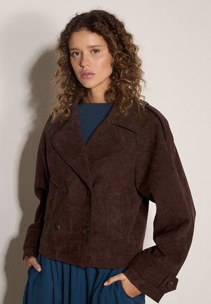 Femme aux cheveux bouclés portant une veste croisée marron foncé sur une robe bleue, mains dans les poches, sur un fond clair uni.