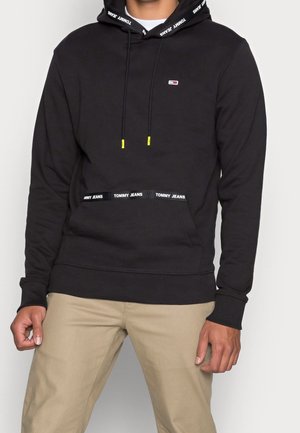 Osoba ubrana w czarną bluzę z kapturem Tommy Jeans z logo na piersi, markową podszewką kaptura i beżowe spodnie, stojąca na jednolitym tle.
