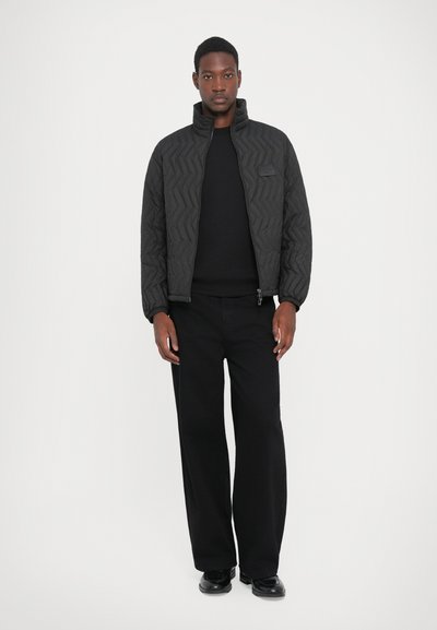Man wearing a black quilted jacket, black sweater, wide-leg black pants, and black shoes standing against a plain white background.