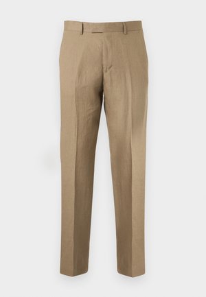 Rövid szőke hajú férfi fekete bőrkabátban, fehér ingben, bézs nadrágban és fekete cipőben, egyszínű háttér előtt áll.