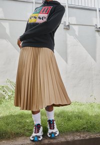 Černá mikina s barevným potiskem na přední straně, skládací béžová sukně, bílé ponožky a vícbarevné tenisky se vyznačují kontrastními texturami a designy.