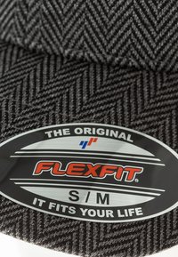Čierna čiapka z látky s vzorom rybacej kosti obsahujúca okrúhlu nálepku s nápisom "FLEXFIT", veľkosť S/M a slogan "FIT PRE VAŠE ŽIVOT."