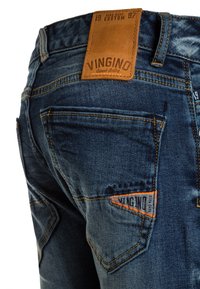 Pantalones vaqueros de denim azul oscuro con un bolsillo trasero que tiene una etiqueta de la marca Vingino cosida en naranja y un parche de cuero marrón en la cintura.