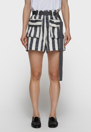 Mujer con pantalones cortos de cintura alta con rayas verticales en blanco y negro, grandes bolsillos delanteros y un cinturón negro que cuelga suelto a un lado.