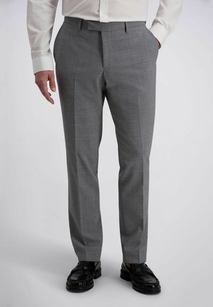 Mann trägt graue Anzughose, schwarze Slipper und ein weißes Langarmhemd, steht mit einer Hand in der Tasche.