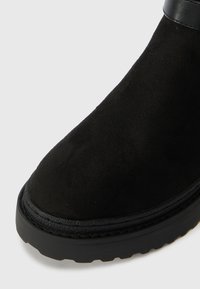 Bota ankle em camurça preta com uma parte superior texturizada, bico arredondado e um detalhe suave em pele preta no tornozelo. Sola grossa com um padrão de frisos.