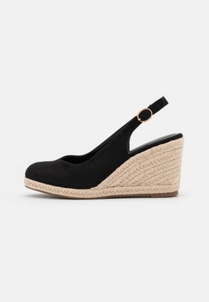Sort kilehælssko med beige flettet espadrille hæl og guldfarvet spænde på rem mod hvid baggrund.