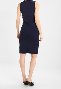 Sinih sininen vartalonmyötäinen mekko, ilman hihoja, polvipituinen, takana halkio. Sujuva pinta ja yksinkertainen siluetti. Mustat korokepohjaiset kengät.