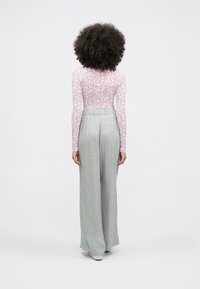 Sieviete ar cirtainiem matiem, tērpusies rozā ziedu raksta garroku krekliņā un vaļīgās gaiši pelēkās ziedu biksēs, stāvot ar muguru pret skatītāju uz vienkrāsaina fona.