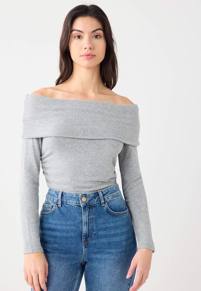 Jeune femme portant un haut gris clair à manches longues et épaules dénudées, et un jean bleu taille haute, debout devant un fond blanc uni.