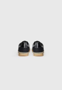 Adidași negri cu partea superioară din fabrică texturată, accente din piele netedă și talpă din cauciuc natural. Prezintă un guler căptușit și detalii de cusătură minime.