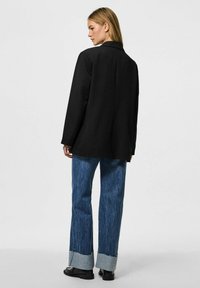 Blazer nero con un design su misura, abbinato a jeans a gamba dritta azzurri con orli sfilacciati e scarpe scure.