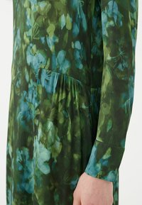 Abito floreale con un motivo verde scuro e blu. Realizzato in tessuto leggero, presenta maniche lunghe e un dettaglio plissé laterale.