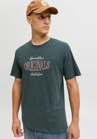 Camiseta verde oscuro de algodón, con un estampado gráfico en blanco y rojo que incluye texto. Combinada con una gorra de béisbol color beige. Corte casual.