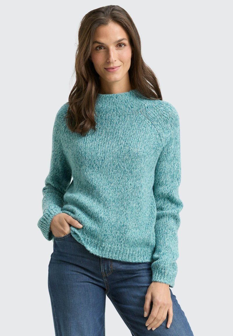 Frau mit langen braunen Haaren, die einen türkisfarbenen Strickpullover und blaue Jeans trägt, posiert mit einer Hand in der Tasche vor einem schlichten Hintergrund.