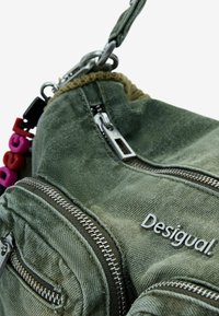 Sac en tissu vert olive avec plusieurs poches zippées, texture douce et quincaillerie argentée. Dispose d'un logo "Desigual!" et d'un charm coloré.
