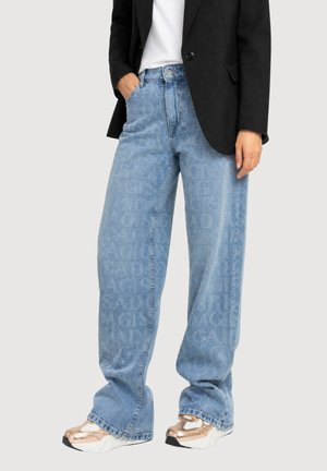 Personne portant un jean bleu clair ample avec un motif de texte subtil, une chemise blanche, un blazer noir et des baskets métalliques rose doré.
