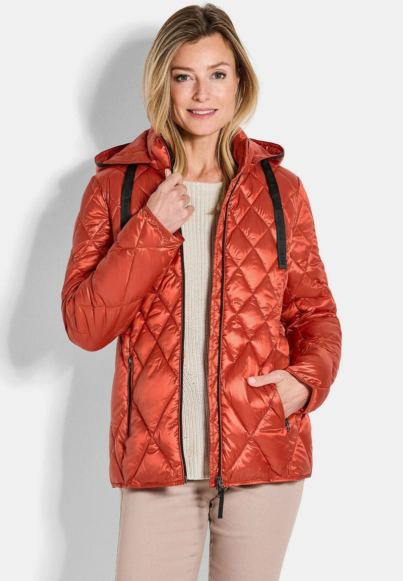 Gesteppte orange Pufferjacke mit Kapuze, ausgestattet mit schwarzen Akzentriemen und Reißverschlusstaschen. Weiche Textur und schlankes, leichtes Design.
