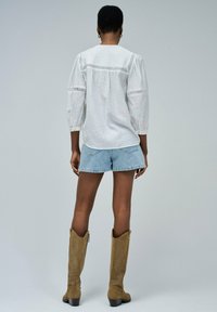 Blusa de campesina blanca con acentos de encaje, mangas tres cuartos y pantalones cortos de mezclilla. La modelo lleva botas de ante color beige hasta la rodilla con un diseño curvado.
