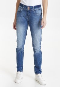 Jeans skinny de denim azul medio, con una cintura de dos botones, cinco bolsillos y una textura desgastada en la parte delantera, combinados con zapatillas blancas.