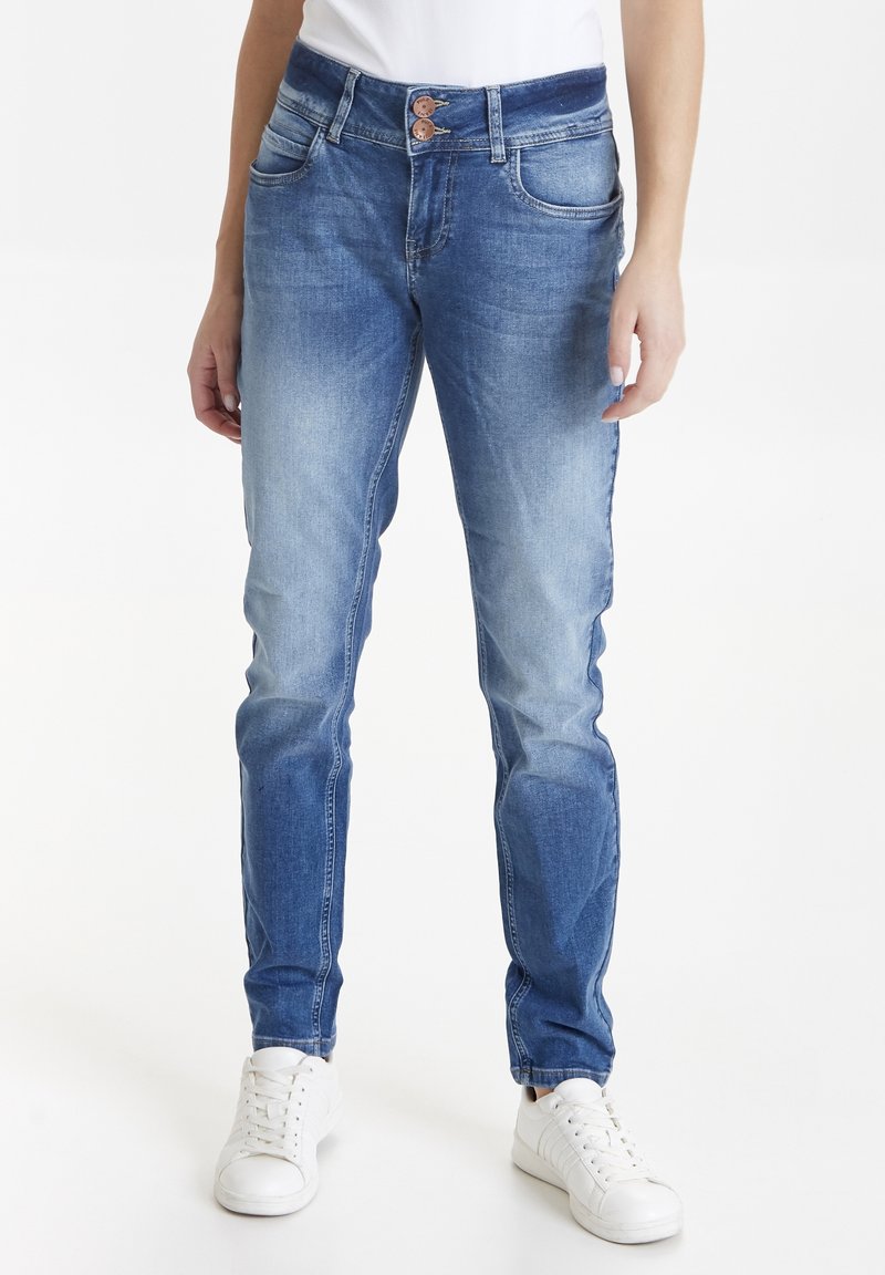 Jeans skinny de denim azul medio, con una cintura de dos botones, cinco bolsillos y una textura desgastada en la parte delantera, combinados con zapatillas blancas.