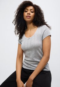 Γκρι κοντομάνικη μπλούζα με στρογγυλή λαιμόκοψη, διακοσμημένη με υφή και δαντέλα. Συνδυασμένη με μαύρο παντελόνι, σε καθιστή θέση.