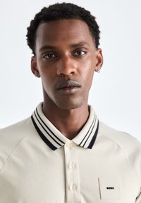 Beige poloshirt met zwart-witte gestreepte kraag, voorzien van een klein vierkant label met "BOSS." Gladde stof, casual ontwerp.