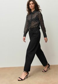 Transparente schwarze Bluse mit weißen Punkten, kombiniert mit hoch taillierten schwarzen Hosen und schwarzen Riemchen-Absätzen.